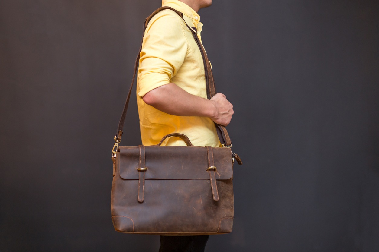 Man carries brown leather messenger bag in the hand on gray background