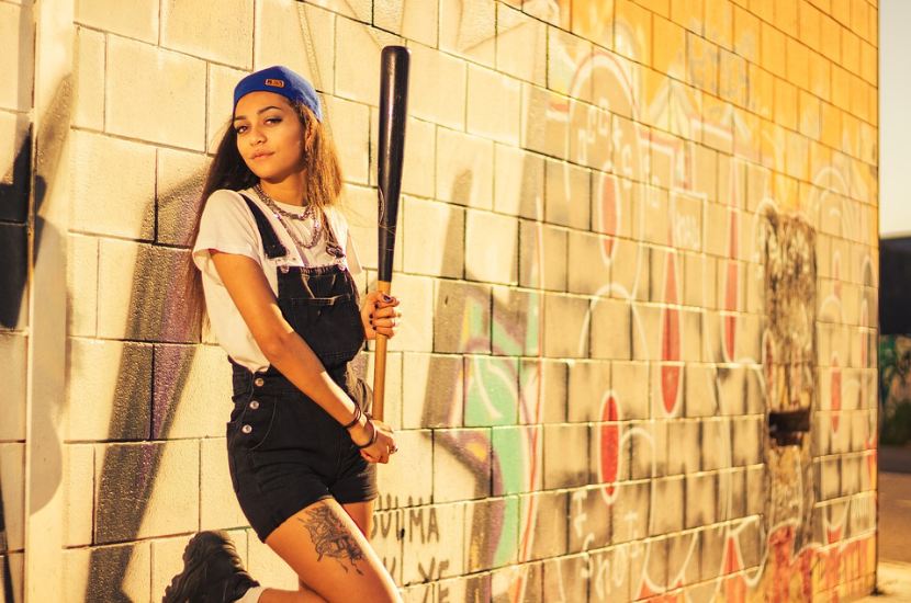 a woman leaning on the wall wearing a baseball game outfit and holding a baseball bat