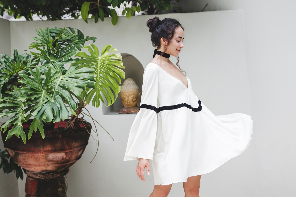 Asian woman with black curly hair enjoying in private villa in Canggu. Wearing white light tunic, barefoot. Casual style, tropical garden. Sunny weather