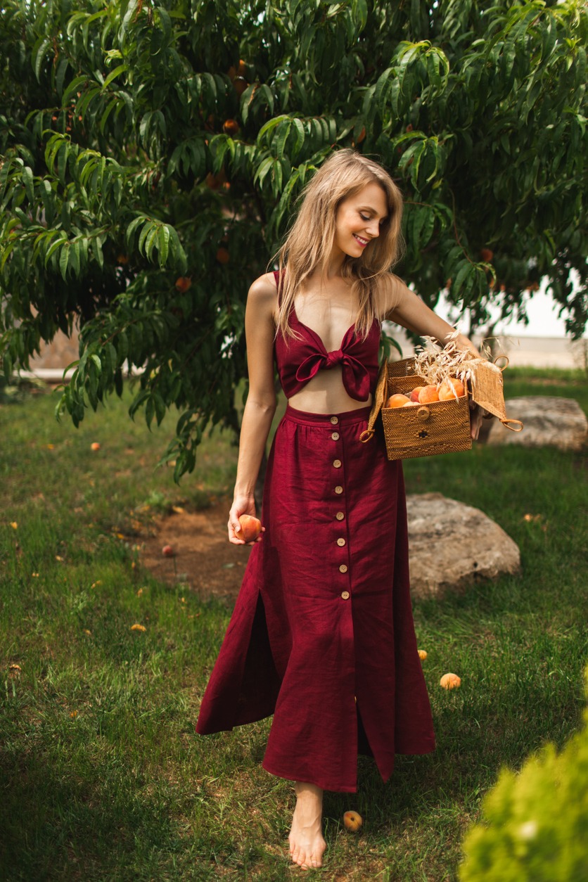 Young beautiful stylish girl in red linen summer dress walking and posing between peach trees. Pretty smile lady with long healthy blonde hair and organic dress walking in home garden. Summer lifestyle concept.