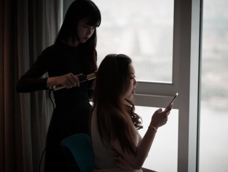 a woman curling another the hair of another woman who’s scrolling through her phone