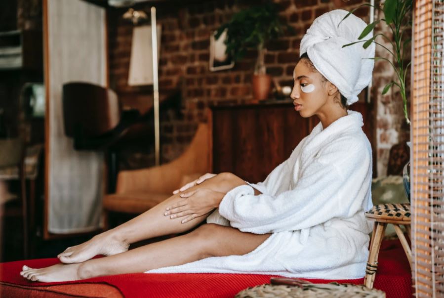 a woman in a bathrobe sitting down and applying cream on her legs