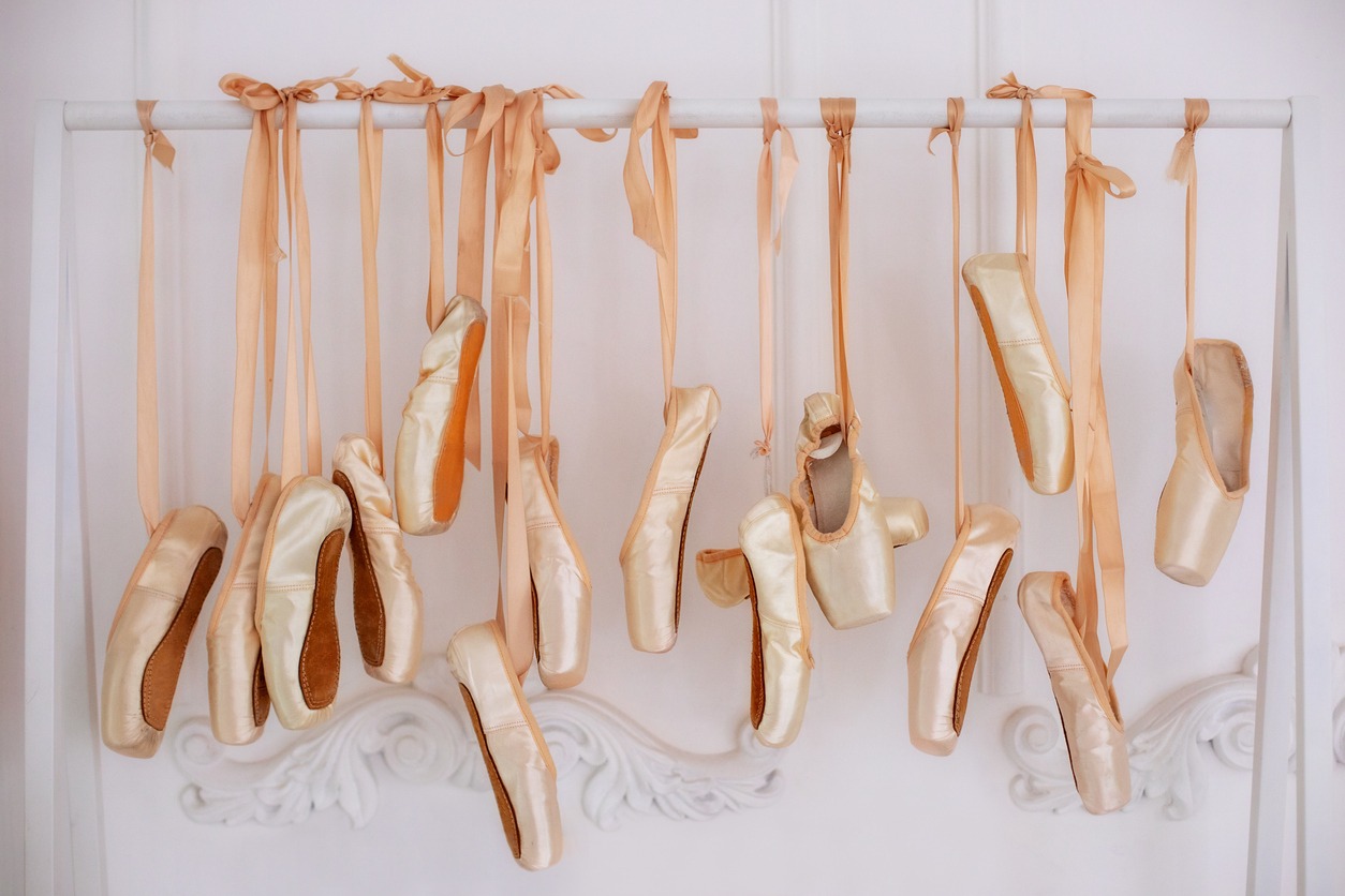 Alt tags: New pointe shoes with satin ribbons hanging on rank. Ballet shoes hang on a bar in the room. Concept of dance, ballet school, ballerinas' clothes. Many hanging ballet shoes on white wall background in a studio