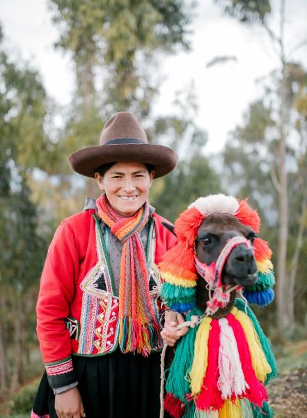 a smiling woman wearing Southwestern clothing and a fringe scarf with a llama with a colorful costume