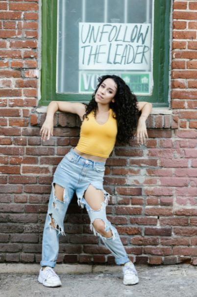 a woman wearing a yellow top and ripped jeans leaning against a brick wall