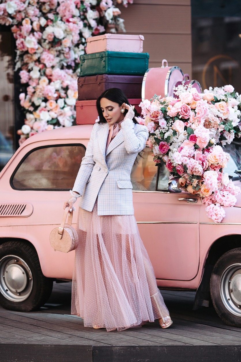 Elegant female fashion model in pink tulle dress and jacket brunette at small retro car with flowers