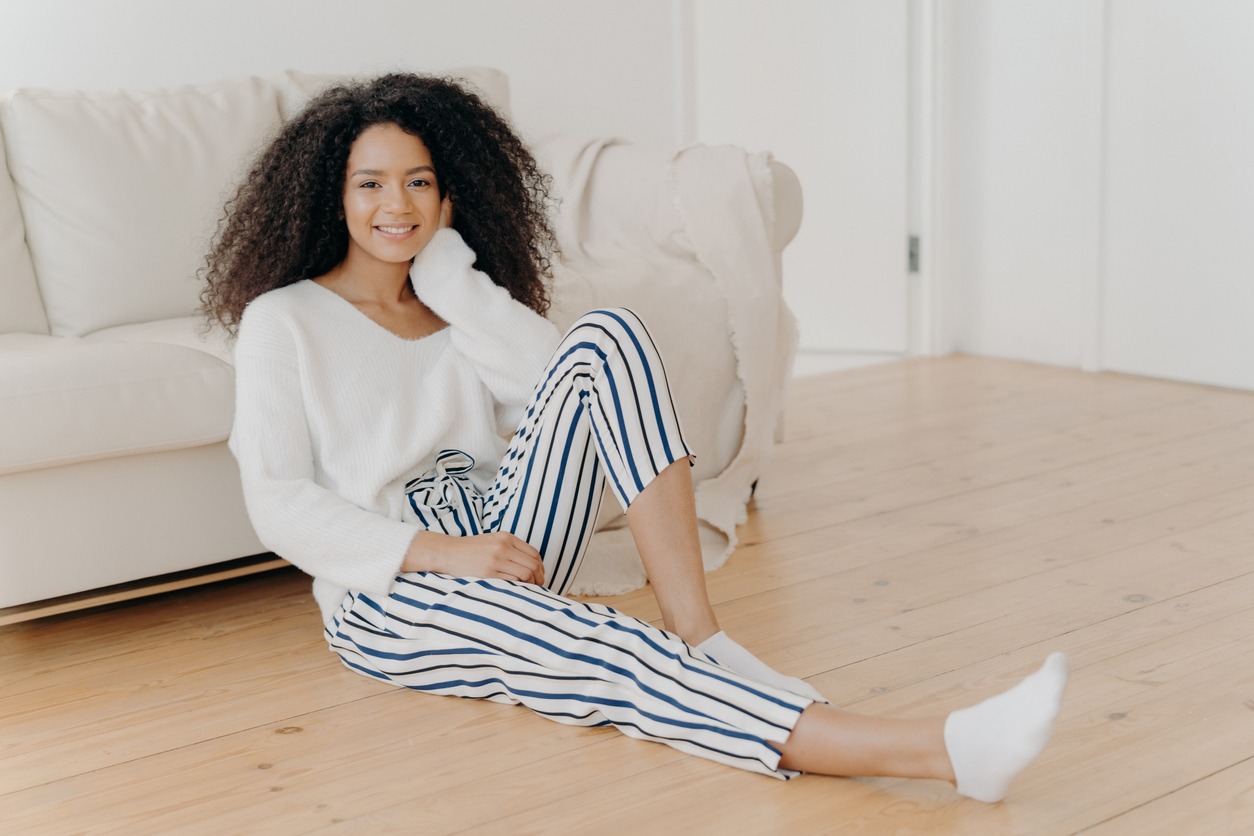 Ethnic girl feels relaxed and satisfied, sits on floor near comfortable sofa in empty room, wears white sweater, striped pants and socks, enjoys domestic atmosphere, enjoys coziness and comfort