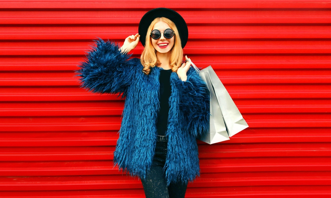 Portrait stylish smiling woman with shopping bags wearing blue faux fur coat, black round hat and sunglasses posing over red wall background