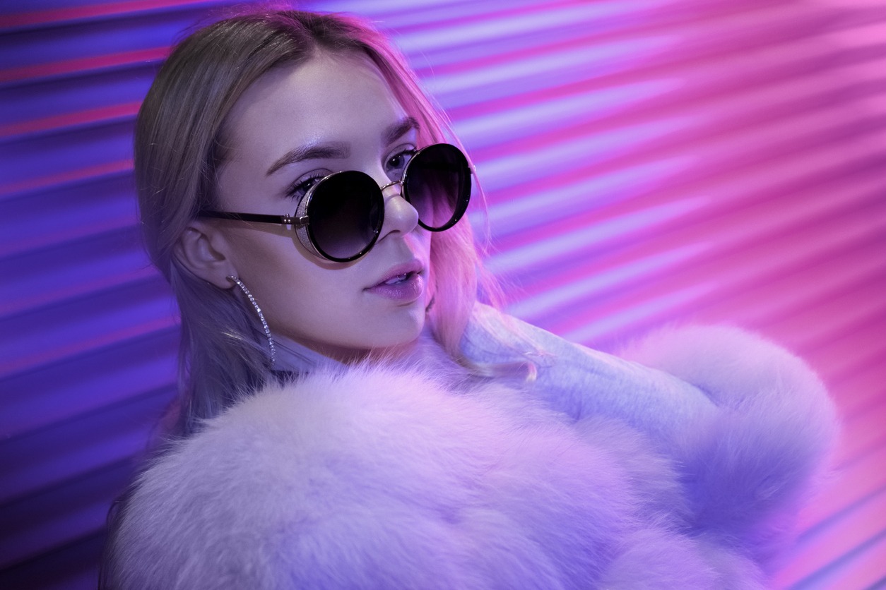 Teen hipster girl in stylish glasses and fur posing on street neon light wall background, female teenager fashion model woman with beautiful face looking at camera in city night light glow, back to 80s, portrait