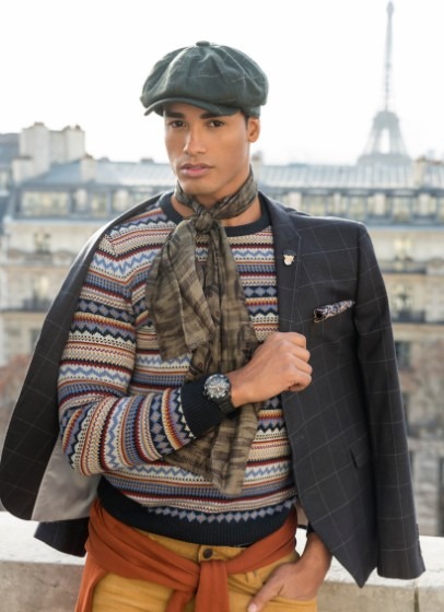 a man wearing a beret hat, scarf, a boho sweater, colorful pants, and a watch with a coat hanging on his shoulder posing in front of a Parisian building