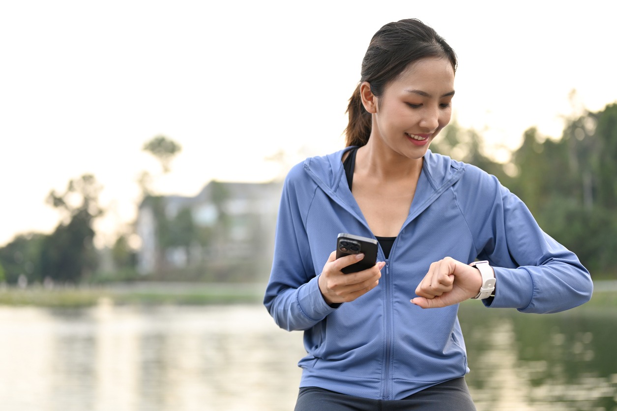 Happy and sporty fit Asian woman in sportswear holding her phone and checking her heart rate and running miles on her smartwatch