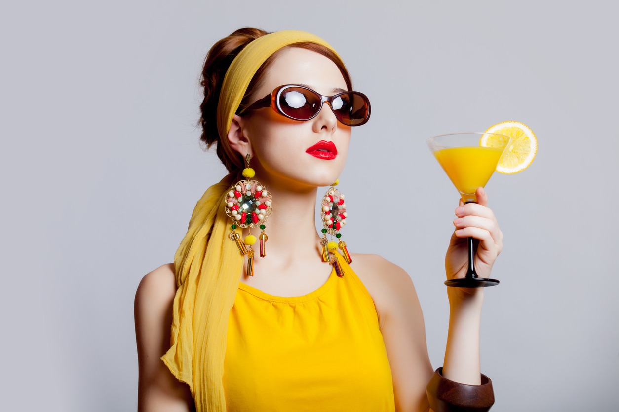 Young redhead girl in 70s clothes style with a tropical cocktail on grey background