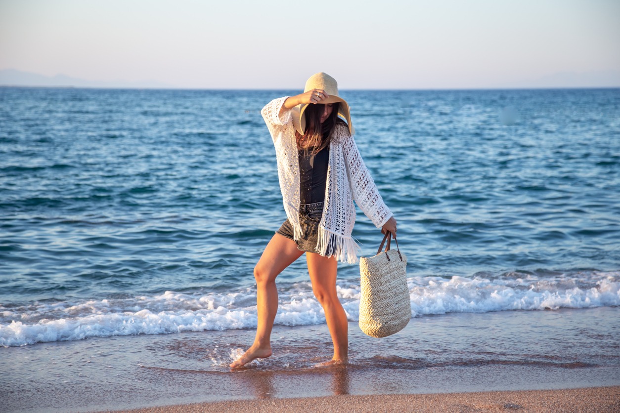 A stunning boho model strolls down the sand while carrying a wicker bag and hat.