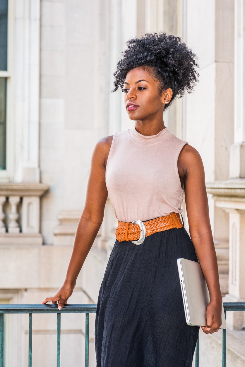 A woman with afro hairstyle wearing sleeveless light color top, belt, black skirt, holding laptop computer, standing in vintage office building.