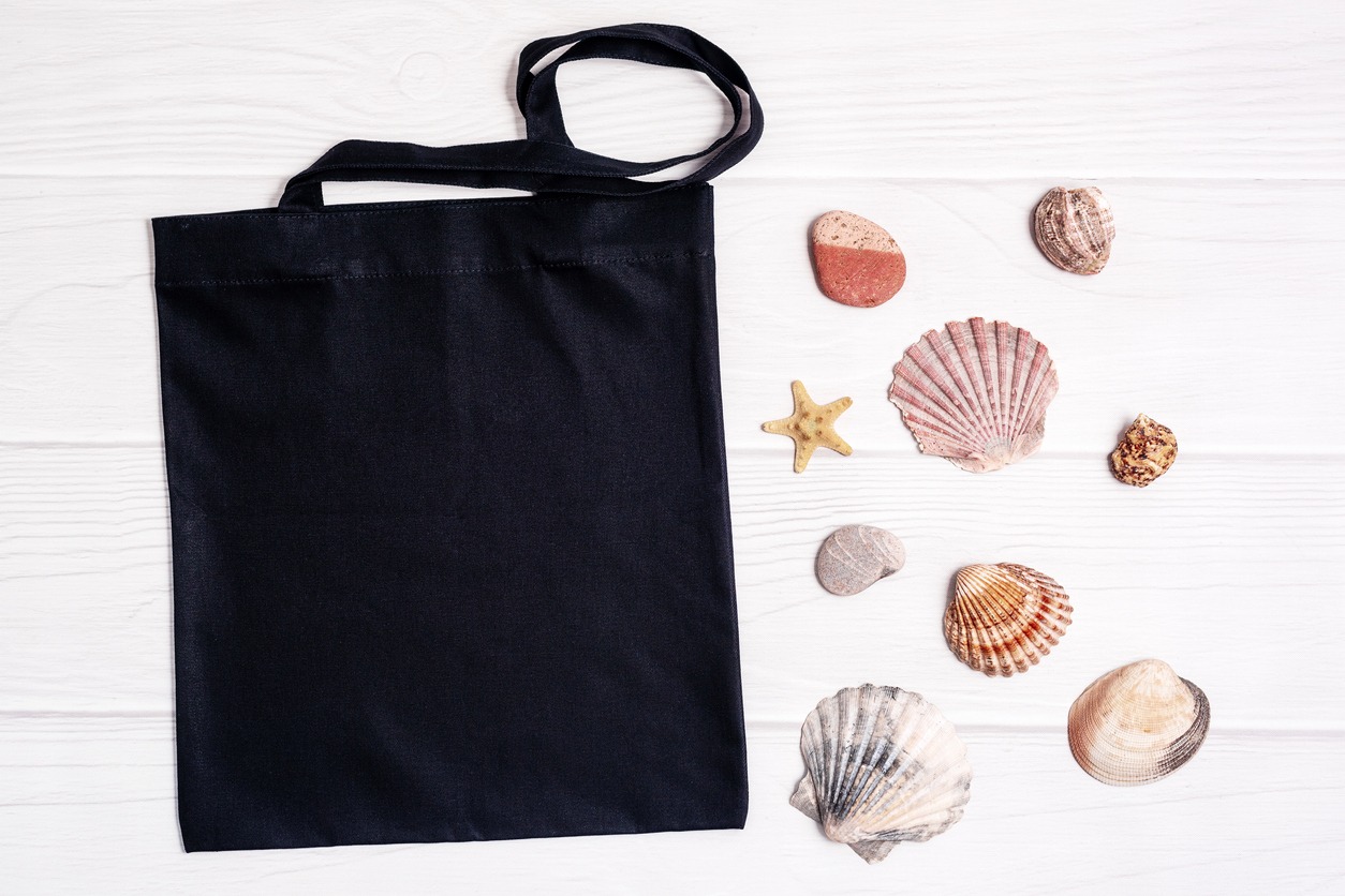 Commercial canvas shopping tote bag with seashells and starfish on a light wood background are displayed on a black blank template.