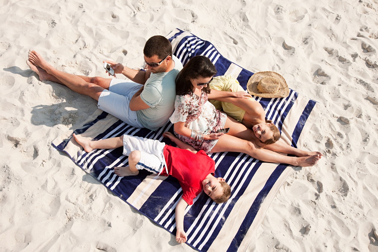 In a big beach towel that is spread out on the sand, a family is seated together.