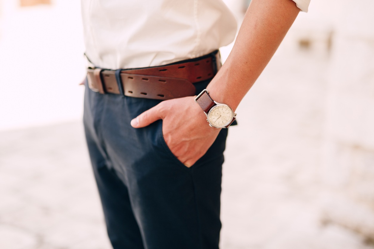 Man in a white shirt with sleeves rolled up, with a watch on his wrist and blue trousers with a brown belt