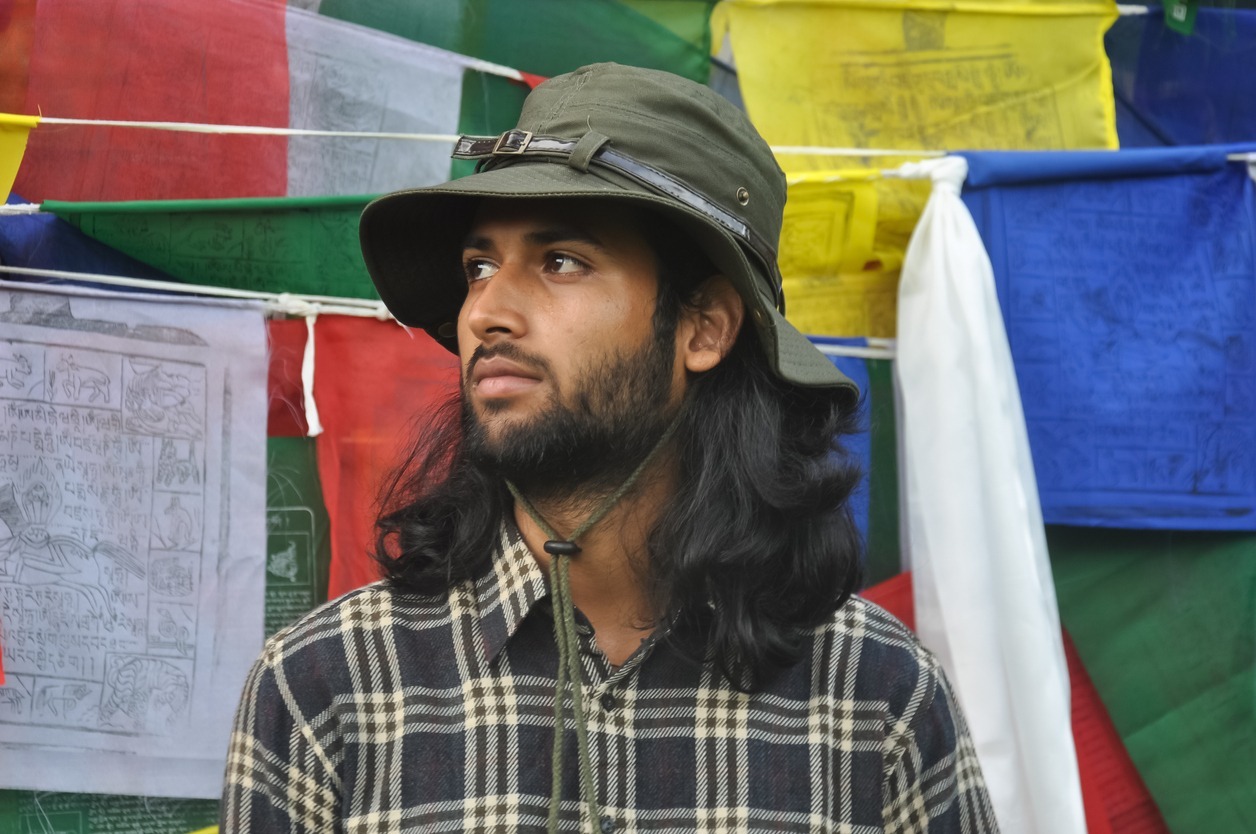 a bearded and long haired male tourist wearing a boonie hat looking sideways while standing against Tibetan prayer flags