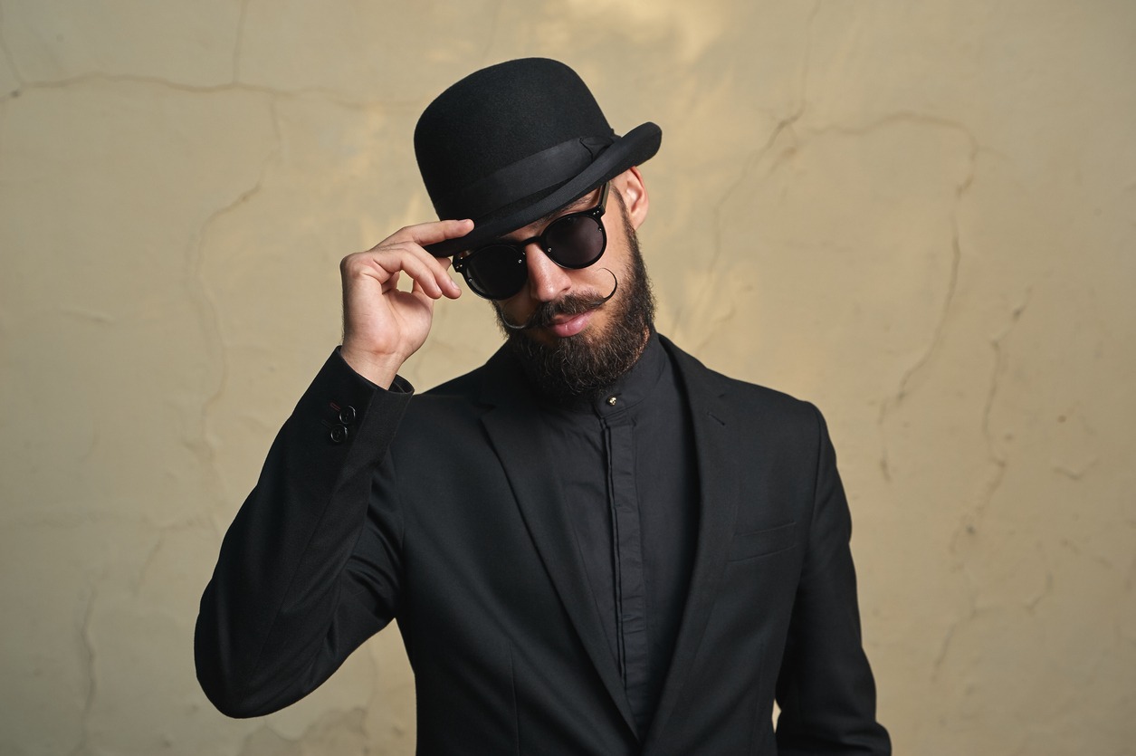 a bearded man in black formal clothing wearing sunglasses and a bolwer hat while holding the hat’s brim