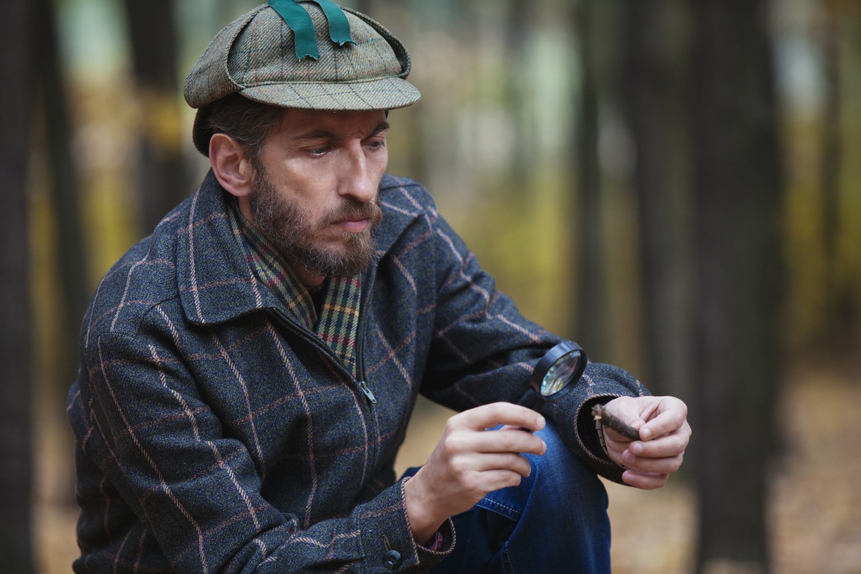 a detective with a beard wearing a deerstalker cap and a plaid jacket looking at a fragment of a tree through a magnifying glass in the forest