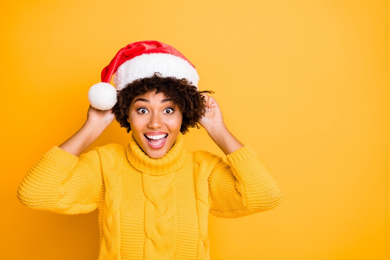 a happy woman with curly hair trying to wear a Santa hat on her head isolated over a yellow color background
