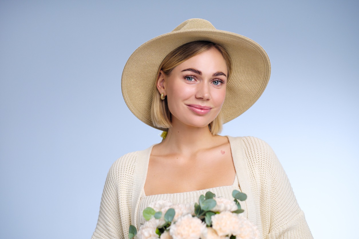 a short haired woman wearing beige outfit and a sun hat holding a bouquet of white flowers on a blue background