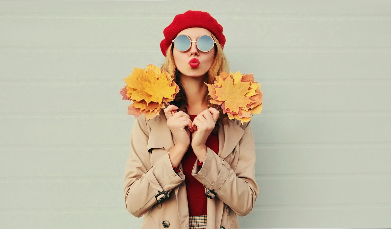 Autumn portrait of beautiful woman with yellow maple leaves, female model blowing red lips sending sweet air kiss wearing french beret over gray background