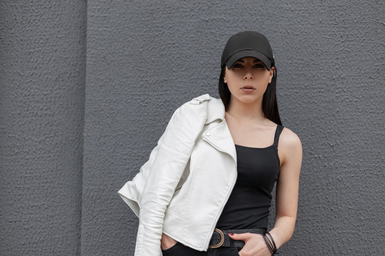 a woman in a black T-shirt, white stylish leather jacket, and black snapback posing near a gray building