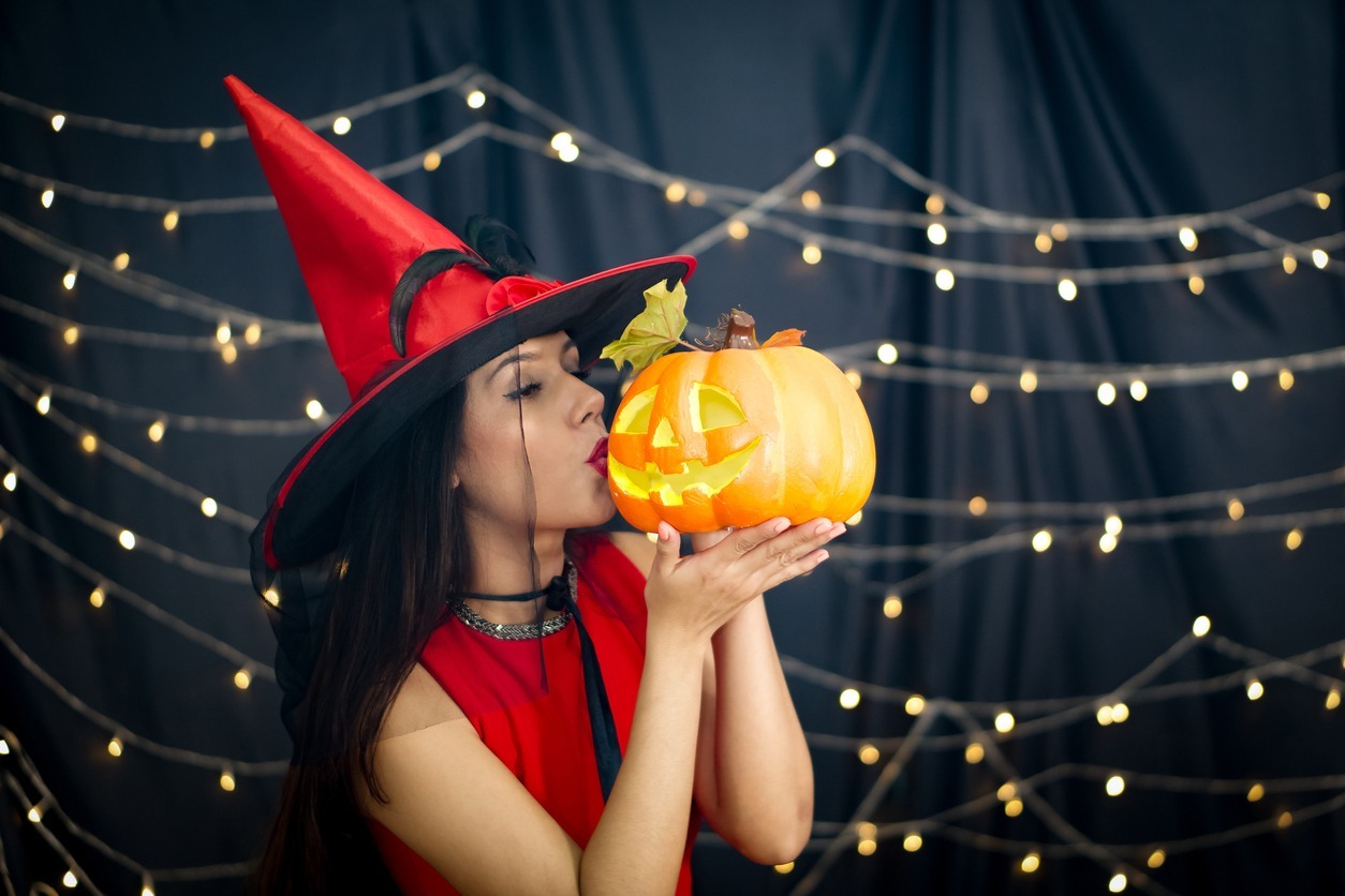 a woman in a red dress wearing a red witch hat and kissing a pumpkin carved in front of a black curtain decoration with lighting