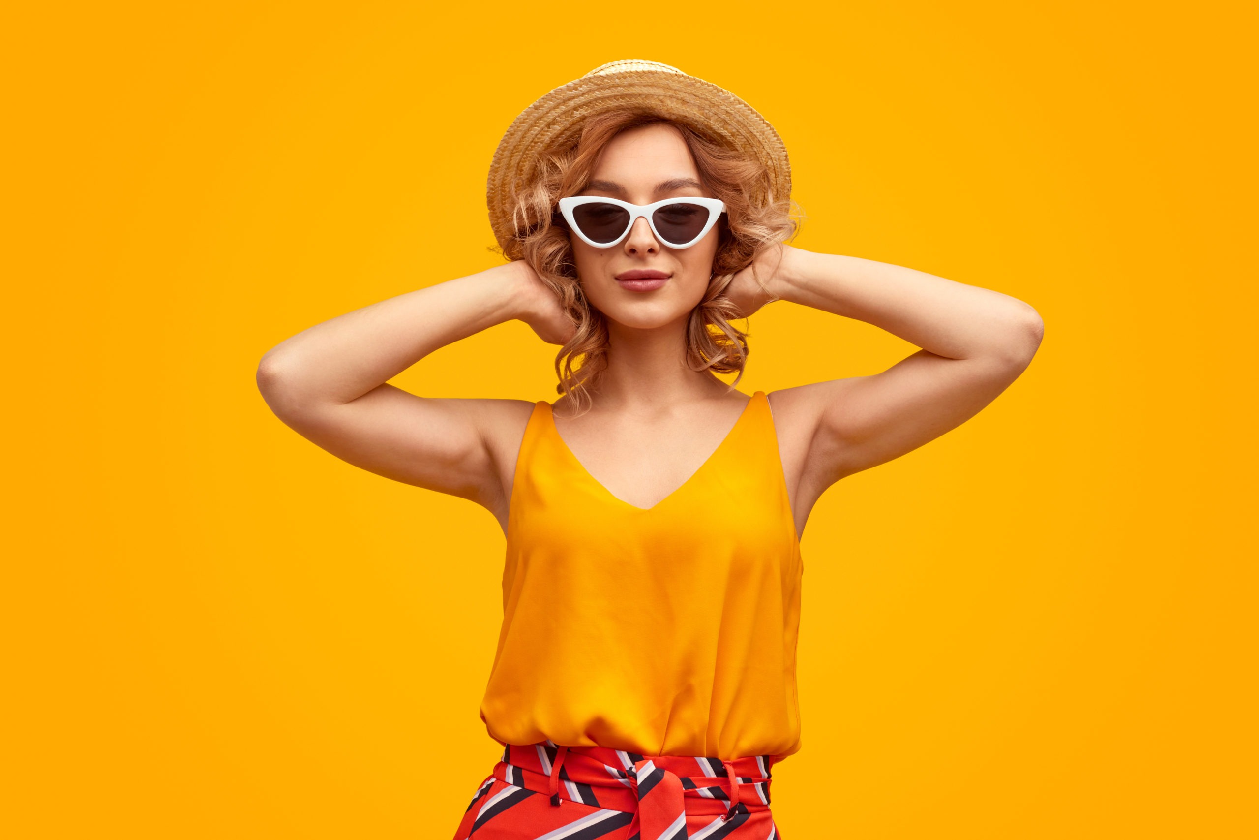 a woman in a stylish outfit and hat looking at the camera and adjusting her wavy blond hair against a vivid yellow background