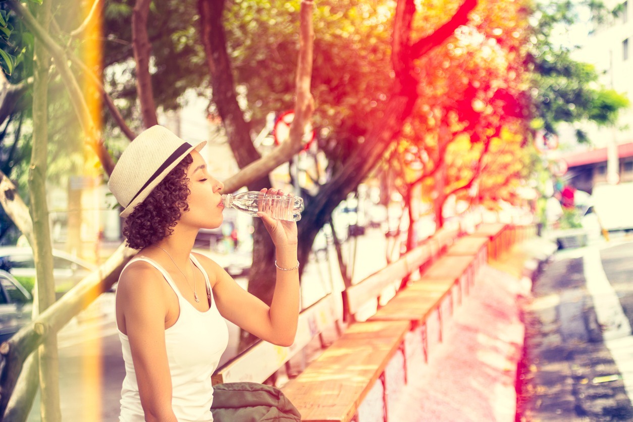 a woman wearing a Panama hat drinking water in a bench at a wooded area protected by a shade