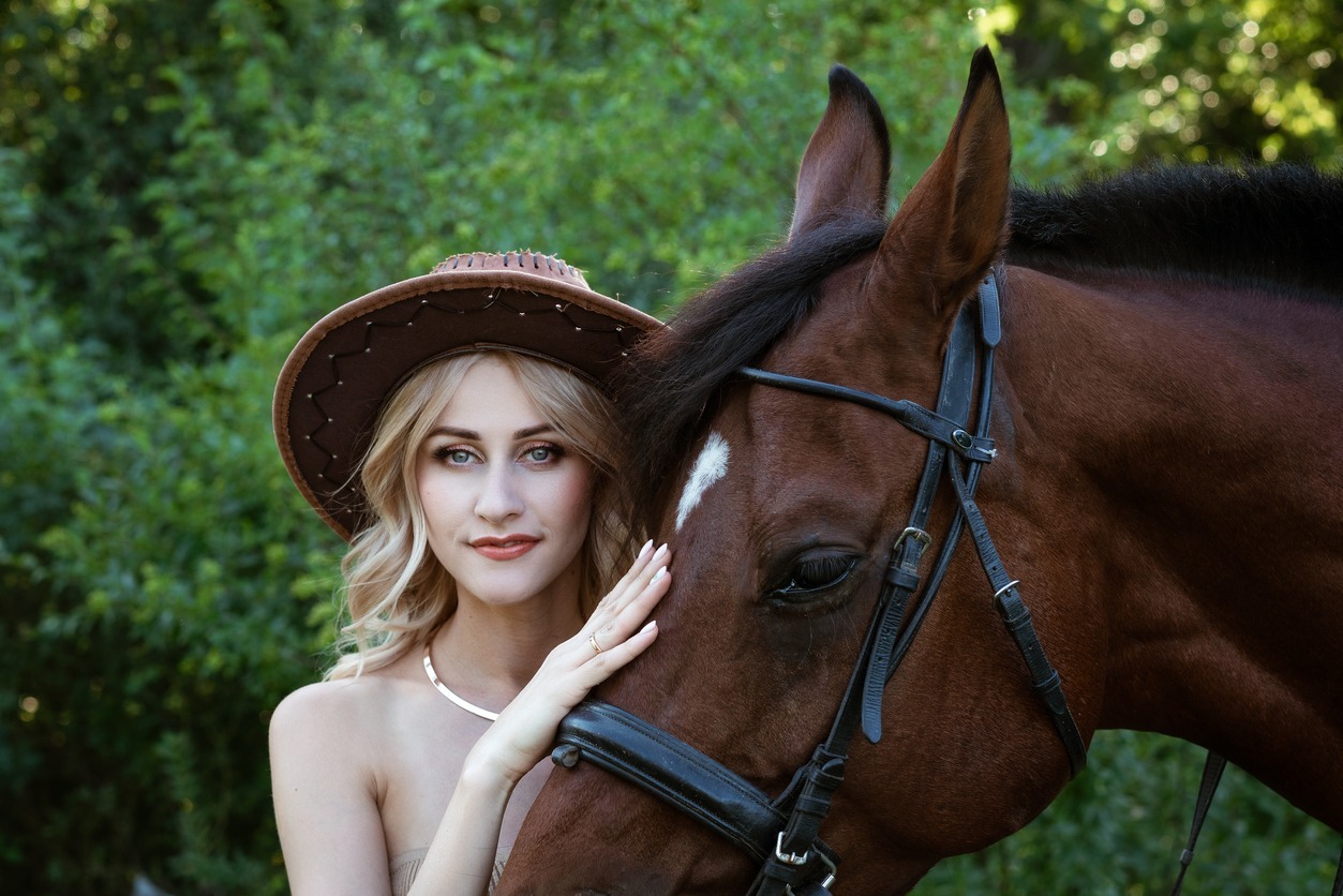 a woman wearing a cowboy hat near and touching a horse