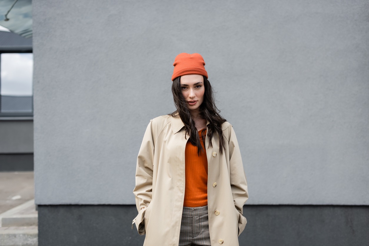 stylish woman in trench coat and beanie hat standing with hands in pockets near building