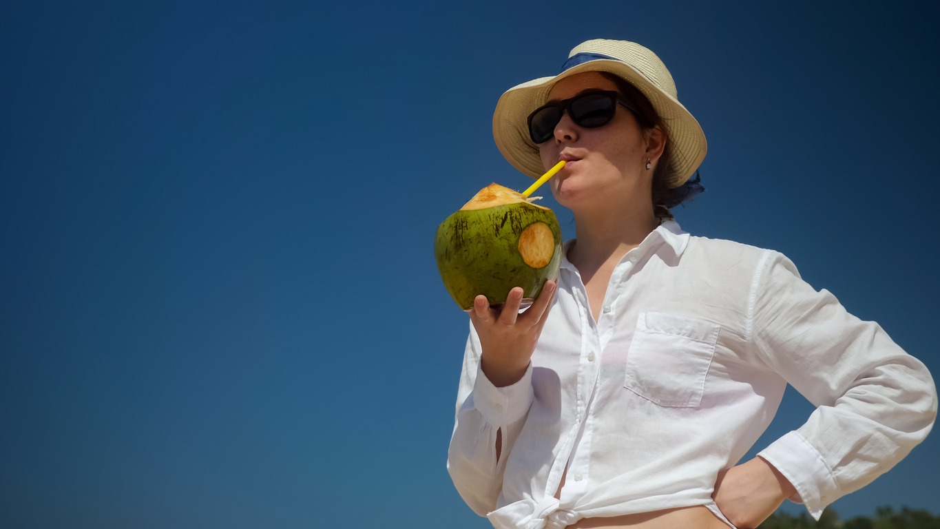 a woman wearing glasses and a Panama hat standing and drinking a coconut with a straw against the blue sky