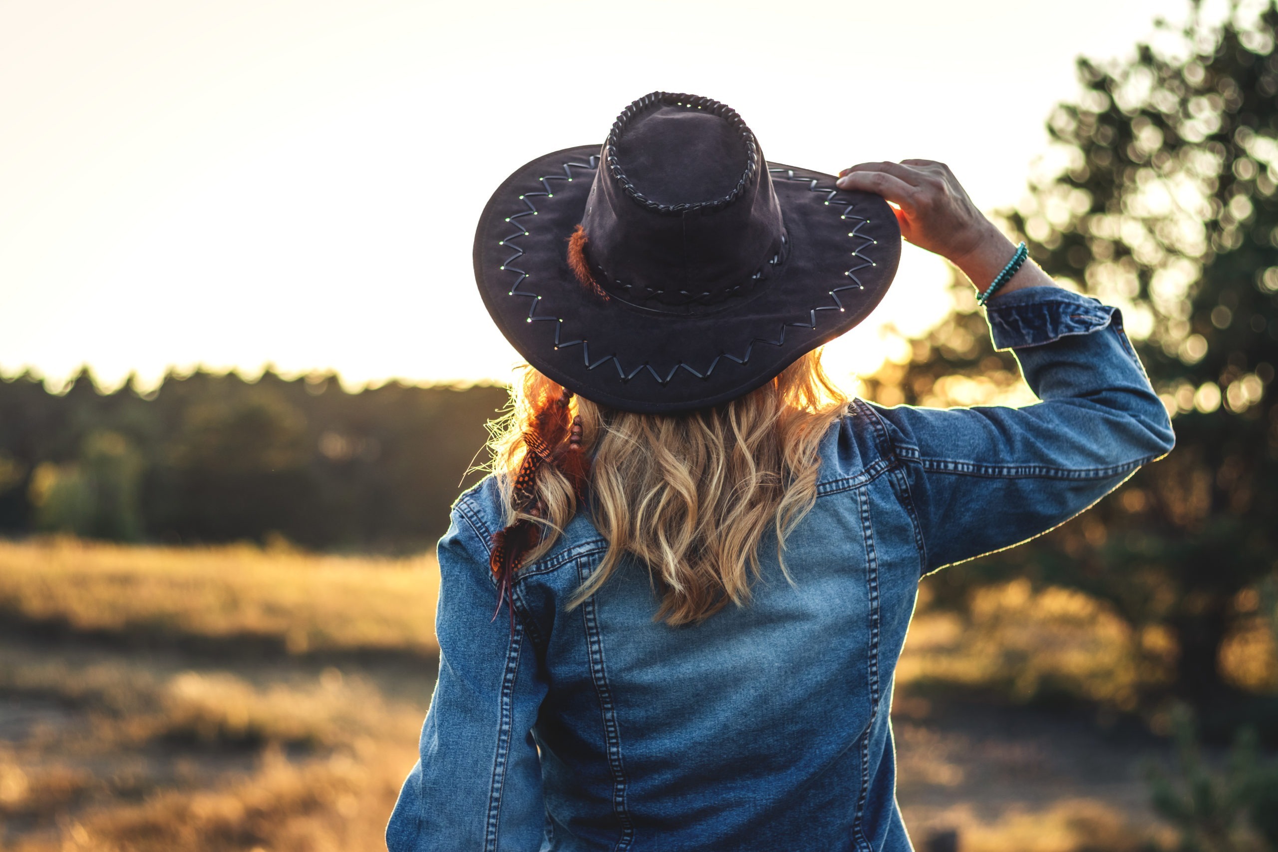 a woman with her back to the camera wearing a cowboy hat and denim jacket enjoying the sunset outdoors