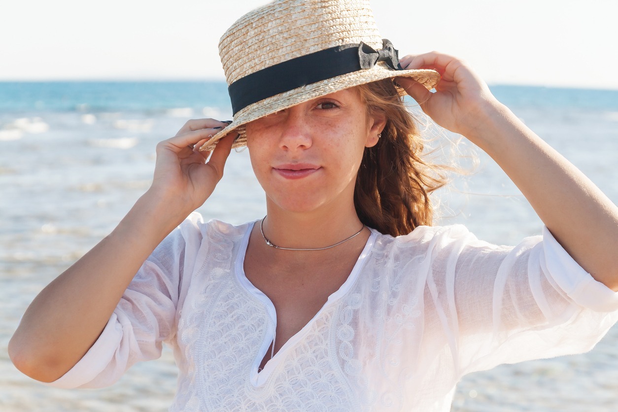 a young woman wearing a lopsided straw hat with the sea at the back