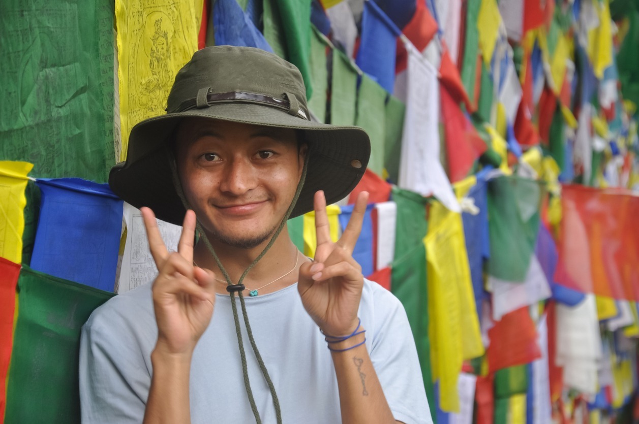 an asian guy wearing a boonie hat looking at the camera and making peace sign with his hands, posing against tibetan buddhist prayer flags