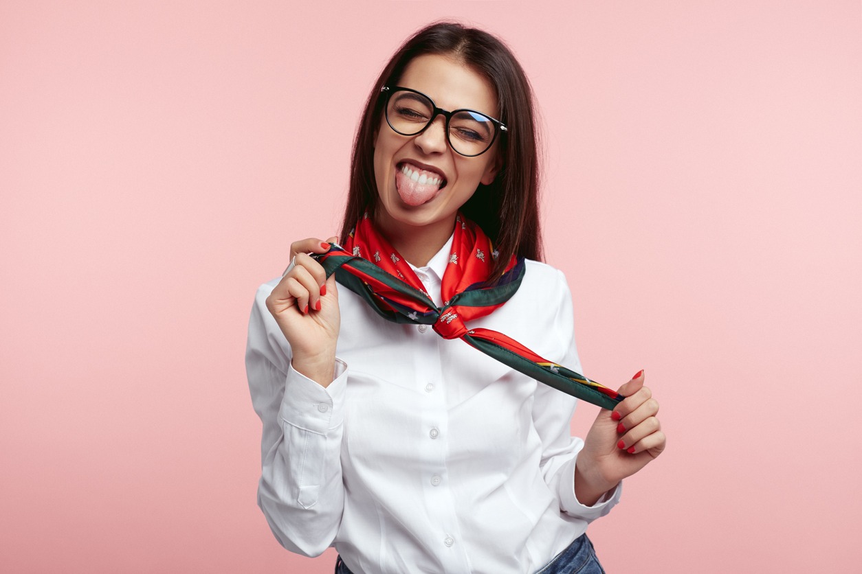 Cheerful pretty lady with eyeglasses, wearing white shirt and holding with her red scarf and showing her tongue with closed eye, standing isolated over a pink background.