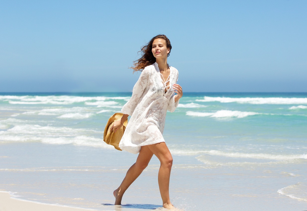 Full length side portrait of a beautiful young woman walking on beach in summer dress