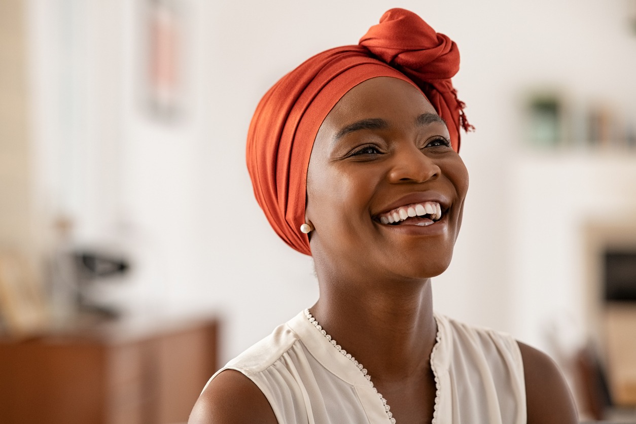 Smiling middle aged African American woman with orange headscarf at home. Beautiful black woman in casual clothing with traditional turban at home laughing. Portrait of mature carefree lady smiling and looking away