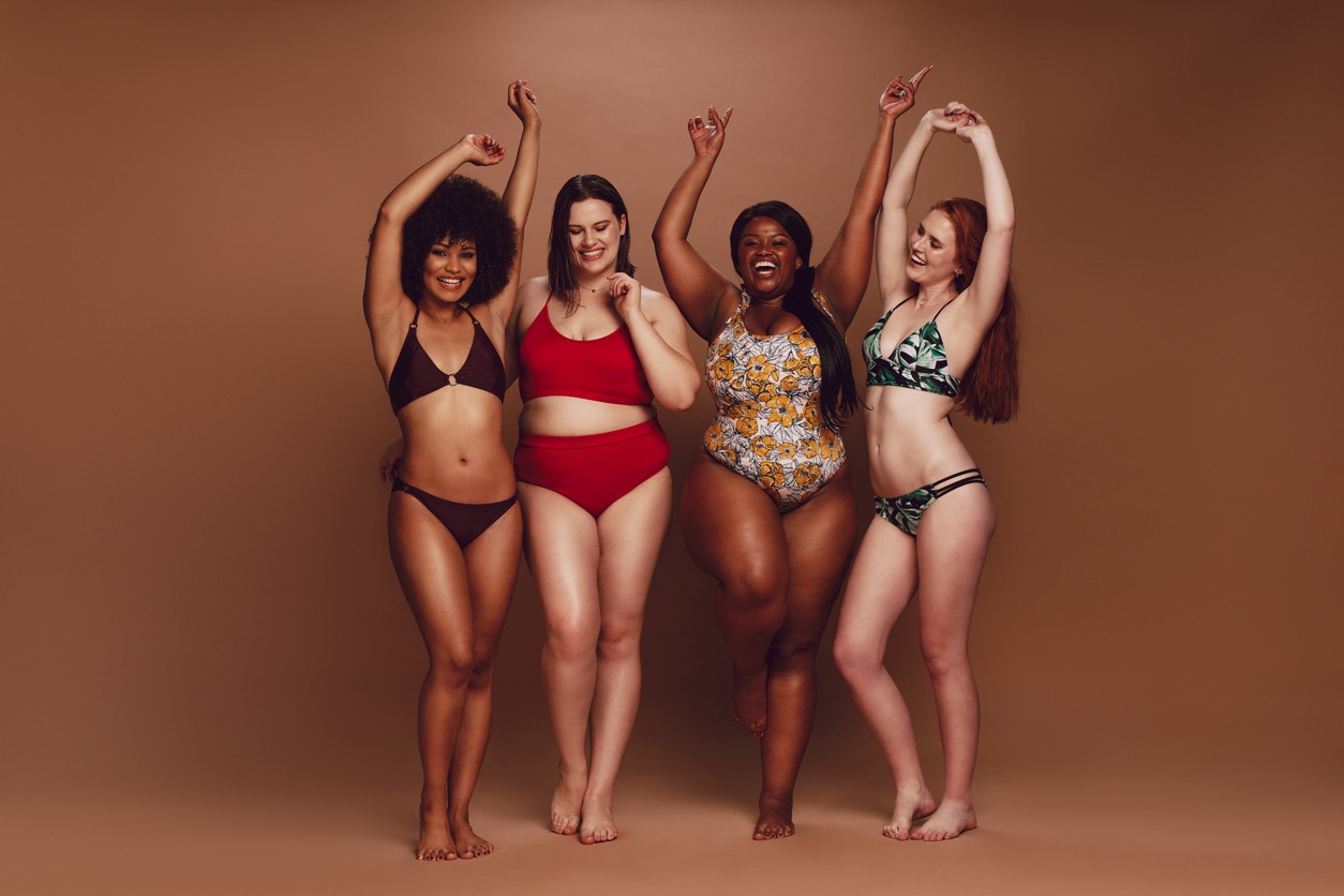 Full length of different size women in bikinis dancing together over brown background. Multi-ethnic women in swimwear enjoying themselves.