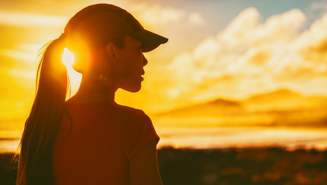 the silhouette of a woman wearing a snapback while walking outside and looking away to the side, sunset