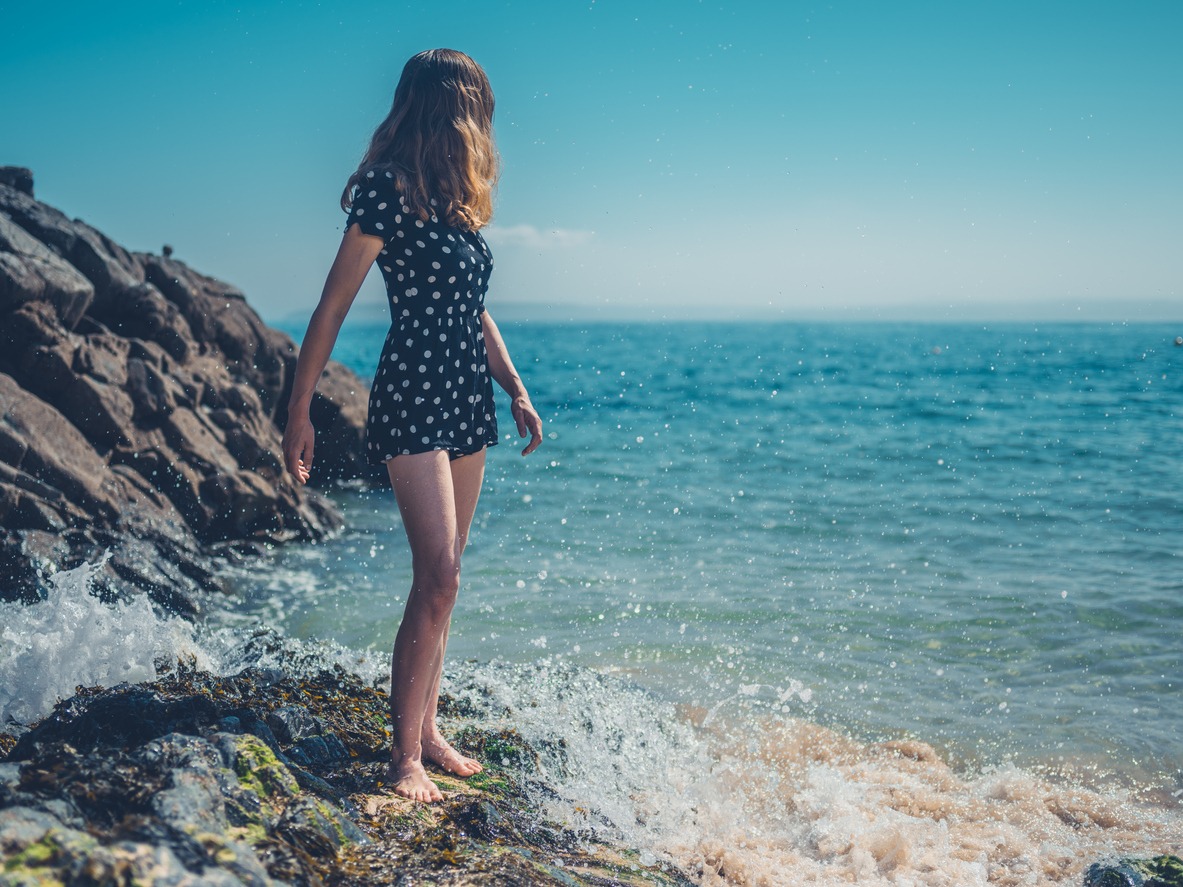A young woman is standing on the rocks of the beach with waves crashing in