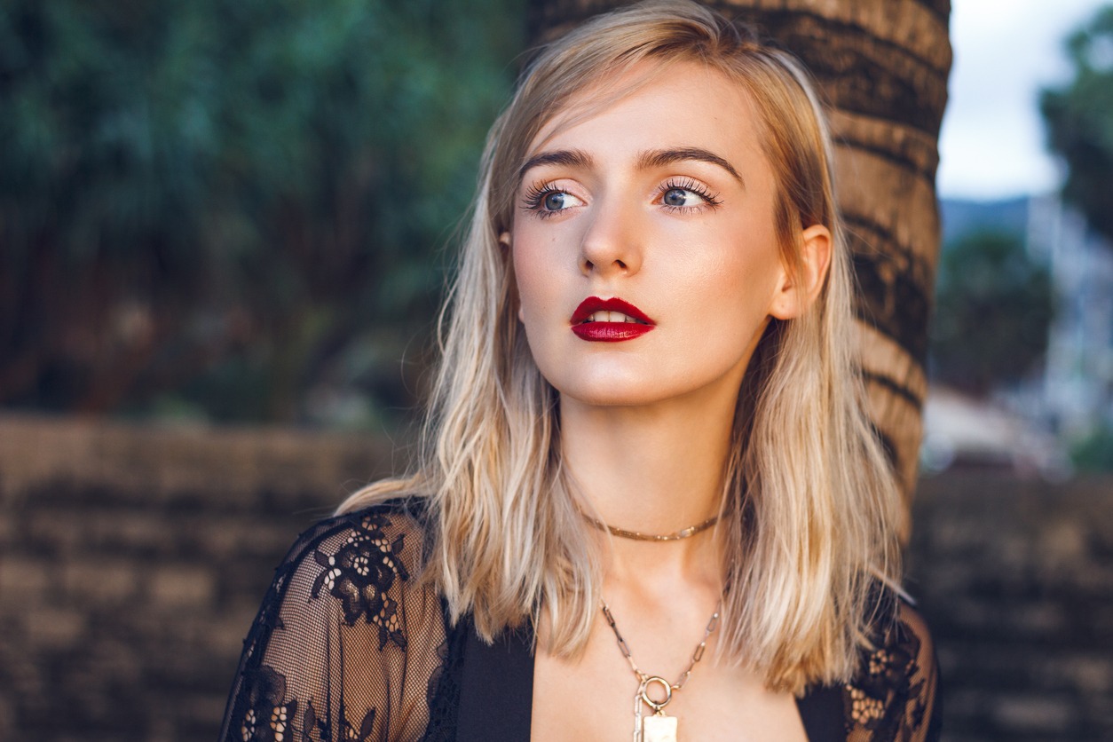 Close-up portrait of a gorgeous young blonde woman with a straight-cut hairstyle, green mysterious eyes, and sexy red lips. She wears fashionable black lace clothing and golden necklaces posing next to the palm