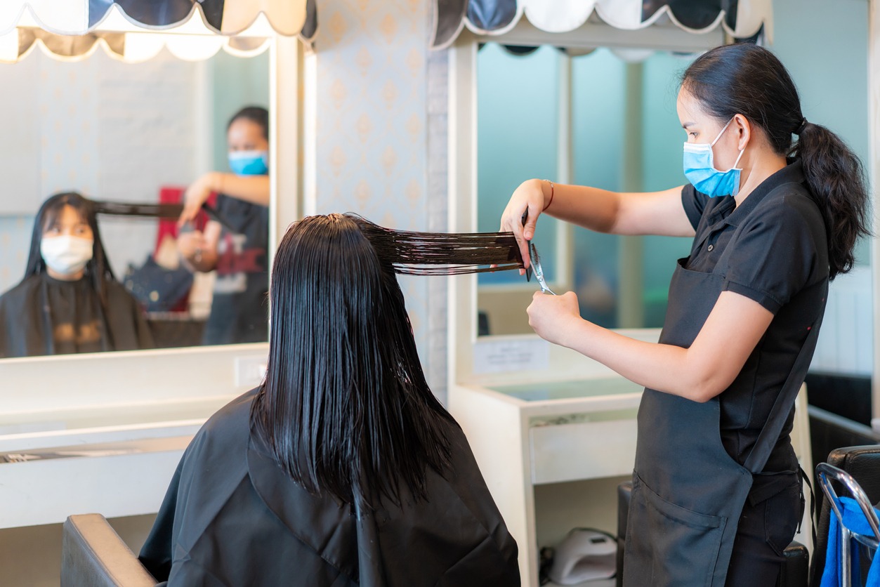Asian young woman wearing face masks to protect themselves from Covid-19 during hairdresser trimming black hair with scissors in a beauty salon