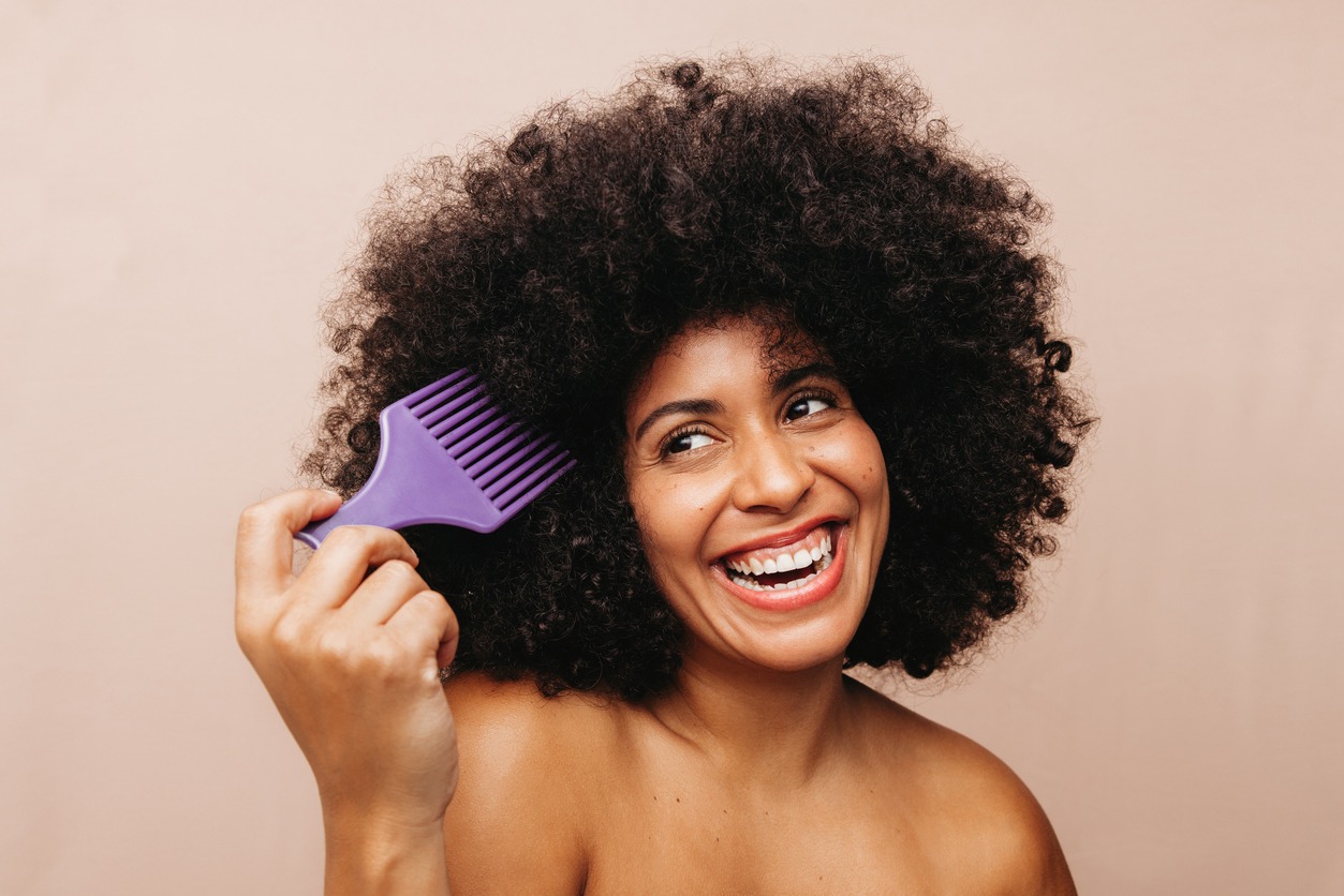Gorgeous woman of color smiling cheerfully while combing her Afro hair. Happy young woman dressing her curly hair with a fork comb. Body-positive woman wearing her natural hair with pride