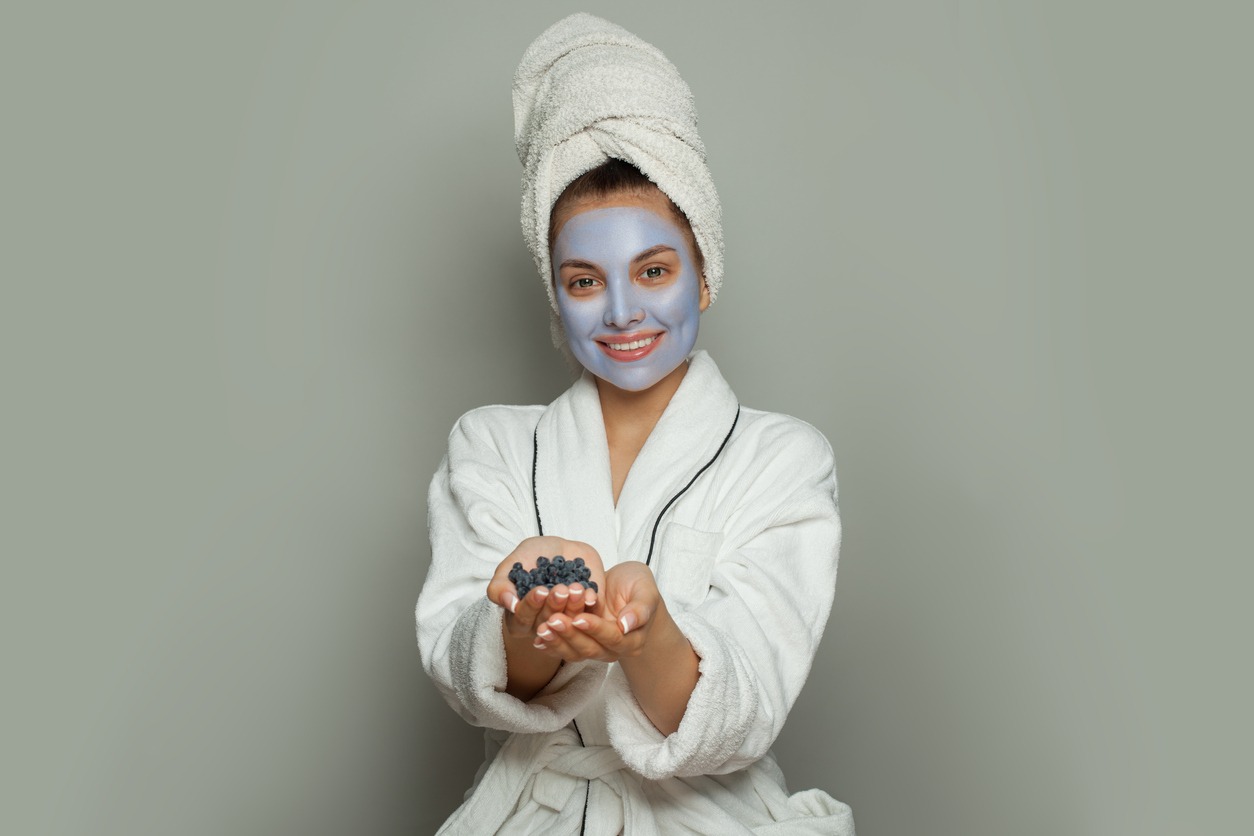 Smiling spa woman in blue cosmetic face mask showing organic blueberries. Facial treatment and skin care concept