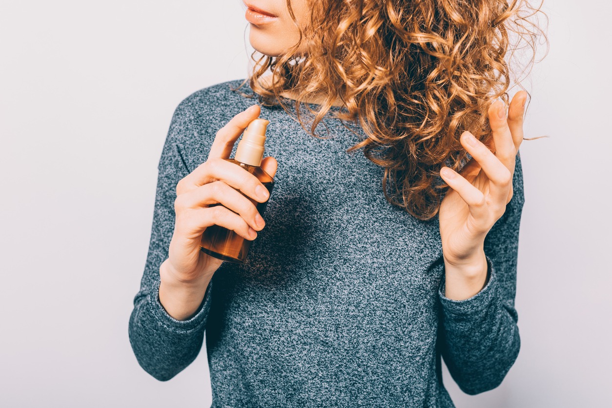 Young woman holding a bottle with nutritional oil applied on her curly brown hair. Female's hands using a cosmetic serum to prevent split ends