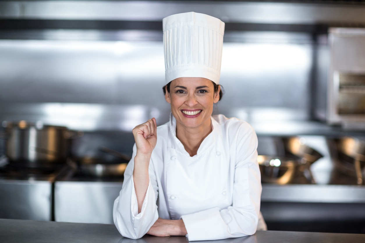 a smiling female chef standing in commercial kitchen and leaning on the counter while wearing her uniform and a chef hat