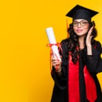 a woman graduate wearing a graduation hat, gown, and eyeglassesholding a diploma isolated a yellow background